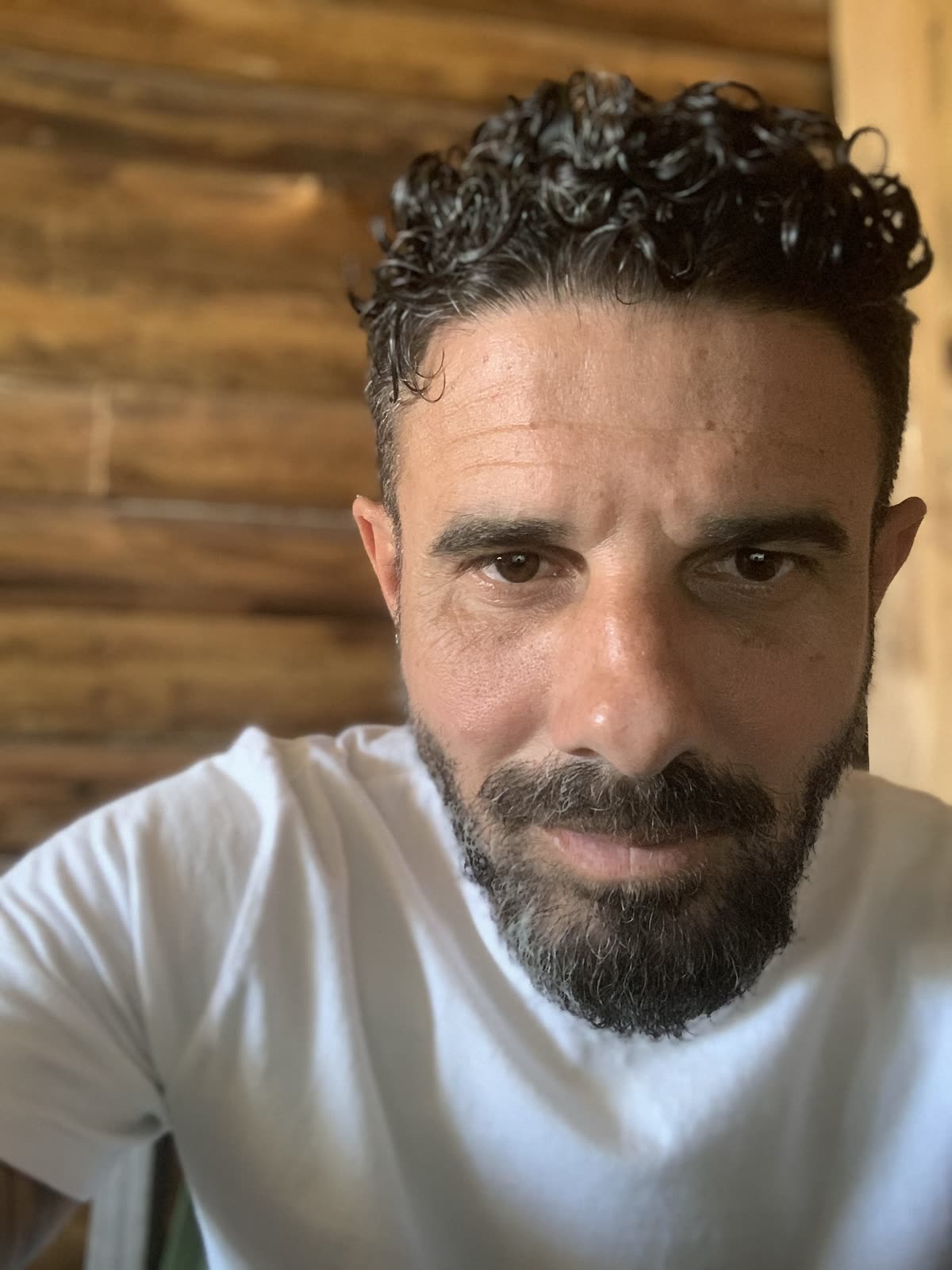 Bruno Callegari olhando direto para a câmera, de camiseta branca, diante de uma parede de madeira