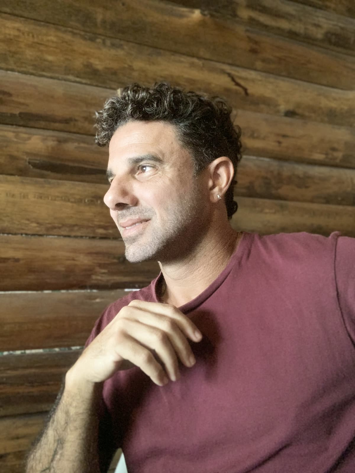 Bruno de perfil, mão no queixo, escutando, de camiseta vinho diante da parede de madeira